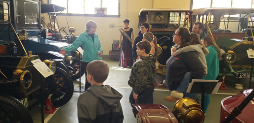 A group of parents, staff and students, engaging in an educational tour of a vintage automobile collection, led by a guide.