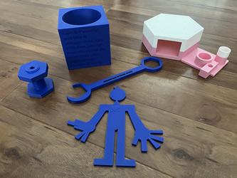 An assortment of vividly colored 3d-printed objects, including a blue wrench and figure, a pink and white structure, and a message stand with an inspiring quote on love and strength.