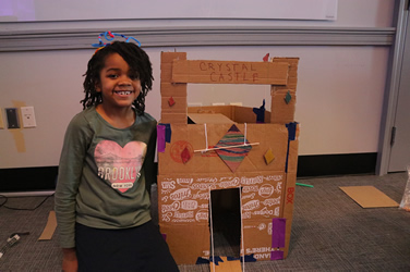 A cheerful young student standing proudly next to her creative project, a cardboard structure labeled 'crystal castle', adorned with colorful decorations.