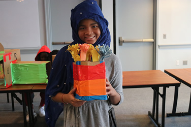 A smiling student at a 'Bring your own Cardboard' event in philadelphia, is pictured holding up a colorful craft project with pride.