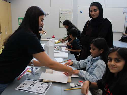 An interactive classroom scene where a teacher is helping young students with an activity while another adult observes.